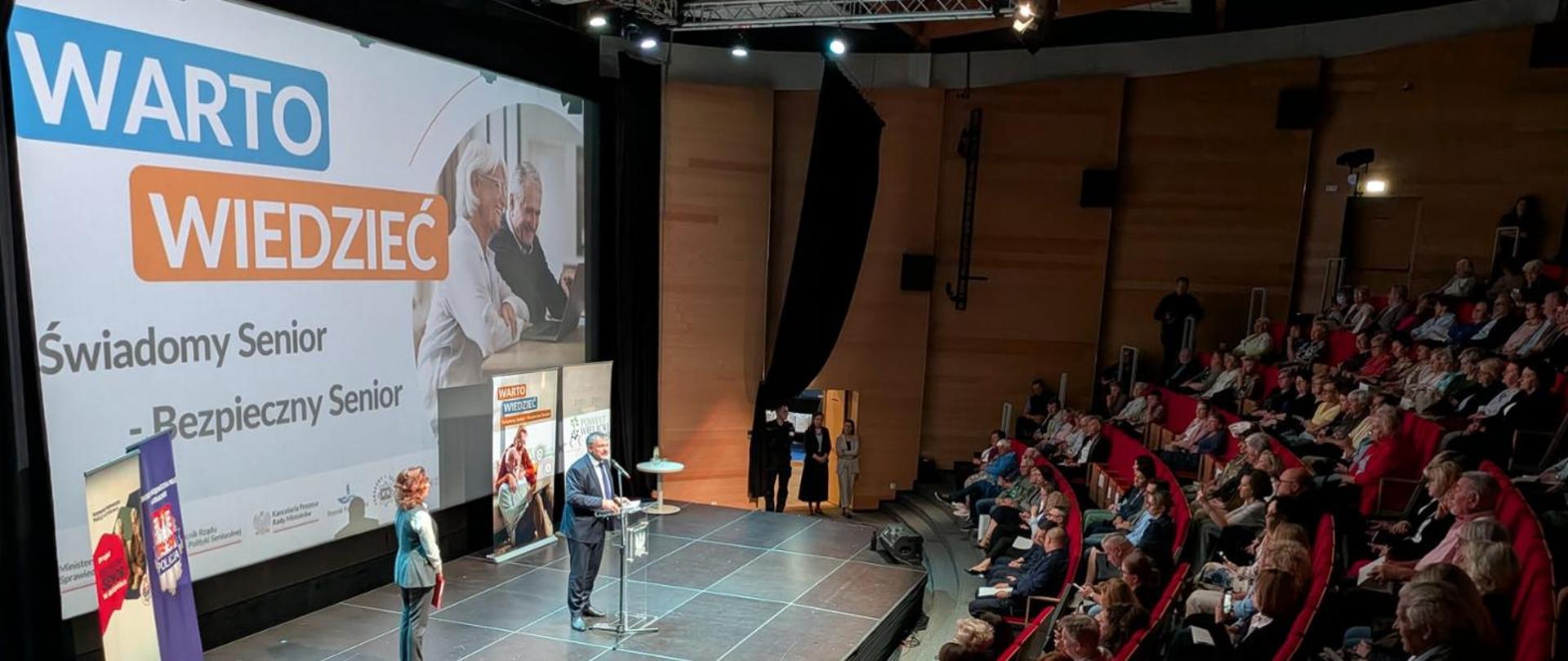 Minister Sprawiedliwości Waldemar Żurek podczas spotkania inaugurującego program edukacji prawnej dla seniorów w Wieliczce oraz prowadząca spotkanie na auli. Po prawej stronie seniorzy i zaproszeni goście.