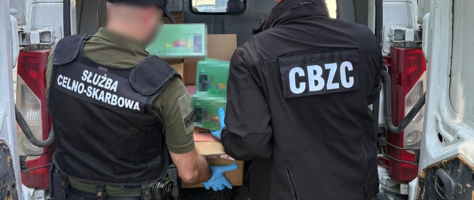 Przy otwartym samochodzie dostawczym stoją dwie osoby, a wewnątrz pojazdu widoczne są kartony i opakowania e-papierosów.
