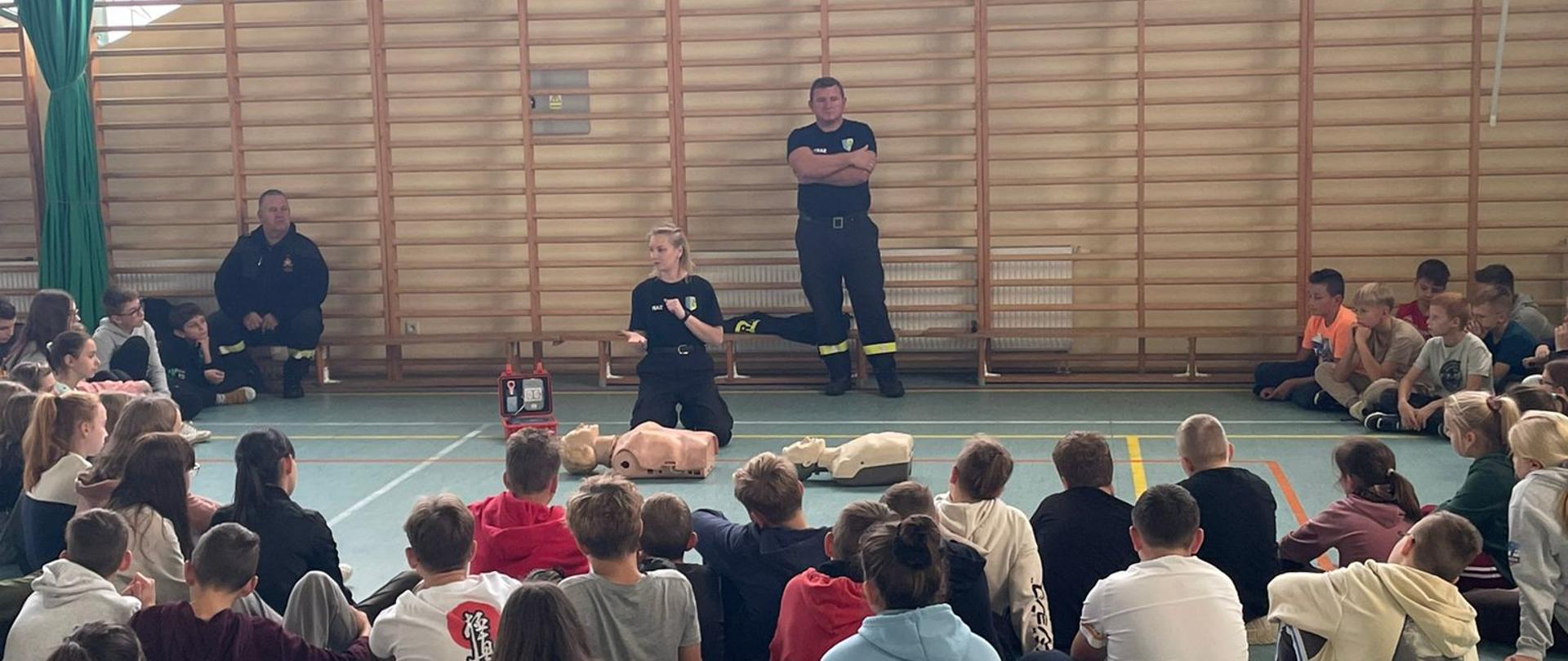 Na zdjęciu znajdują si dzieci siedzące na sali gimnastycznej oraz strażacy, którzy tłumaczą zasady udzielania KPP