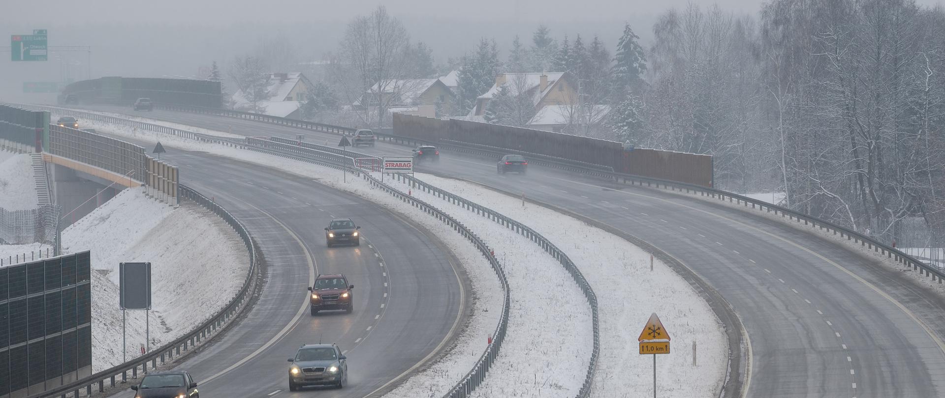 Na zdjęciu widoczne samochody poruszające się drogą ekspresowa S17. Zdjęcie wykonane zimą - odśnieżona droga i niewielka warstwa śniegu pokrywająca pasy zieleni oddzielające jezdnie.