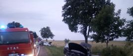 Na pierwszym planie widać rozbite auto osobowe znajdujące się w rowie, obok na jezdni stoi pojazd pożarniczy na sygnałach świetlnych, w tle znajduje sie drugie auto w rowie i samochód strażacki.
