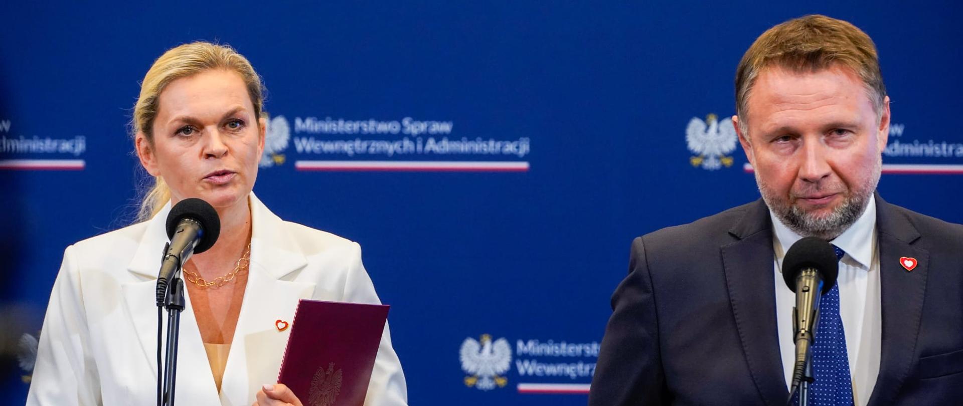 Kobieta i mężczyzna stoją obok siebie na konferencji prasowej. Przed nimi ustawione są mikrofony. Kobieta jest w białym garniturze i trzyma czerwoną teczkę, a mężczyzna jest w granatowym garniturze z krawatem w kropki. W tle widoczny jest baner z godłem Polski i napisem „Ministerstwo Spraw Wewnętrznych i Administracji”.