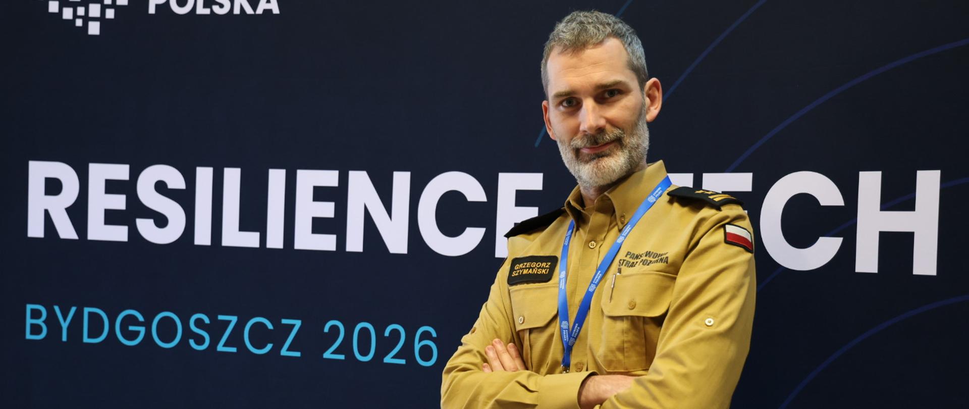 Strażak w mundurze służbowym, z założonymi rękami stoi na tle granatowej ścianki reklamowej z białym i niebieskim napisem. Na szyli ma niebieską smycz firmową.