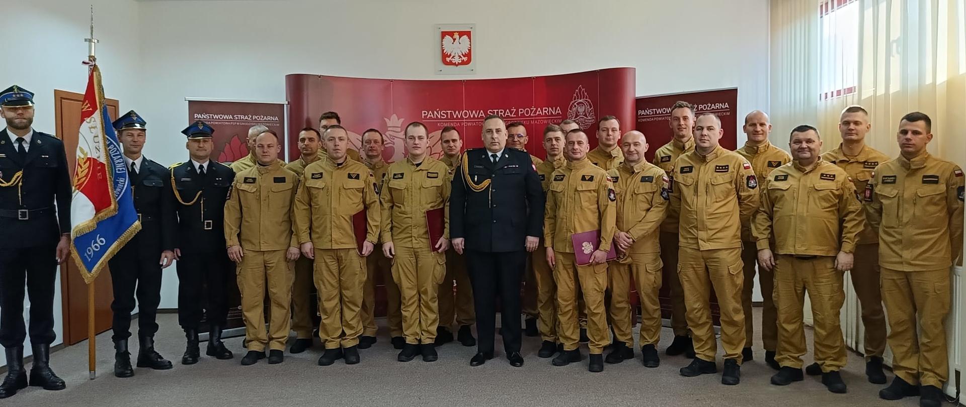 Zdjęcie przedstawia liczną grupę strażaków pozujących razem w sali recepcyjnej. W centralnym punkcie stoi oficer w ciemnogranatowym mundurze wyjściowym ze złotymi sznurami galowymi. Otacza go kilkunastu strażaków ubranych w piaskowe mundury służbowe; niektórzy z nich trzymają w dłoniach pamiątkowe, bordowe foldery z dokumentami.
Po lewej stronie kadru stoi poczet sztandarowy z dumnie prezentowanym sztandarem jednostki. Wszyscy uczestnicy zbiórki są zwróceni przodem do obiektywu. Tło stanowi czerwona ścianka ekspozycyjna z napisem „PAŃSTWOWA STRAŻ POŻARNA” oraz polskie godło wiszące na białej ścianie powyżej. Sala jest jasna, oświetlona światłem dziennym wpadającym przez okno z prawej strony.