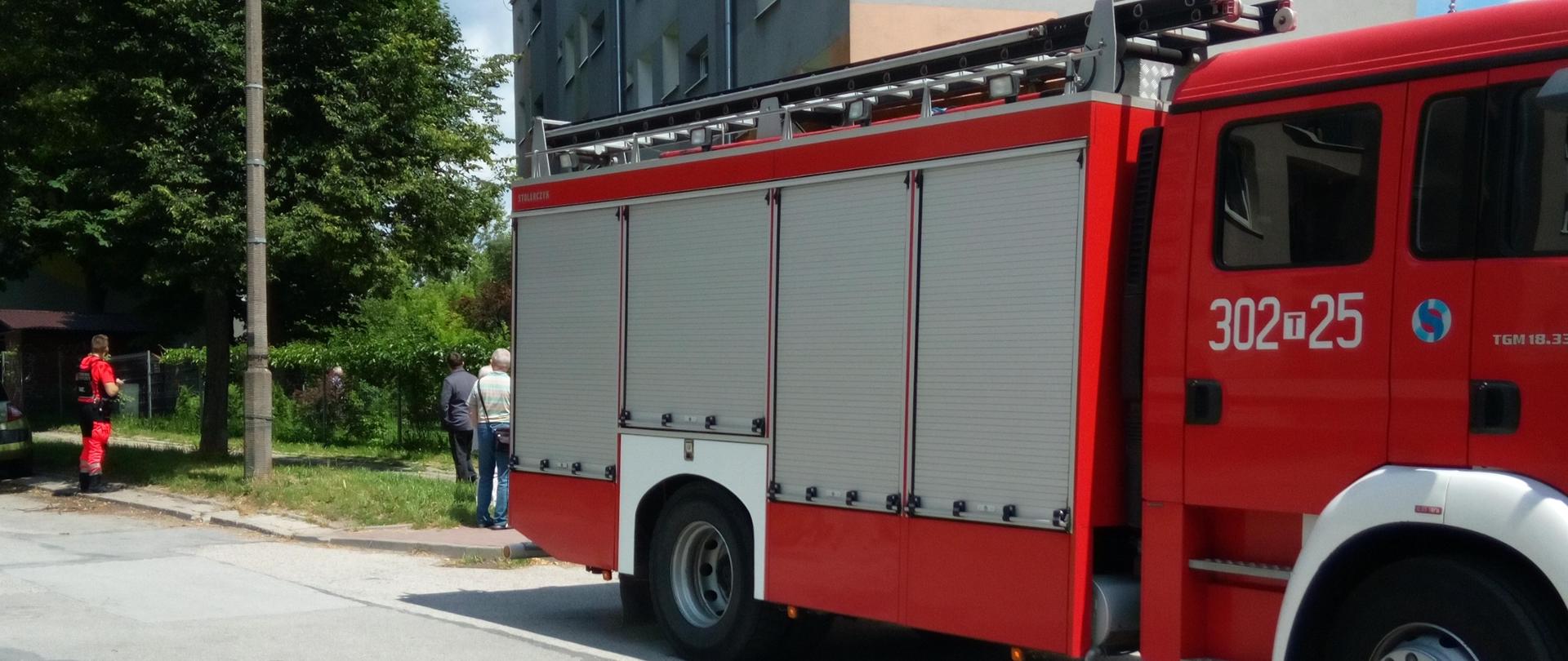 Fotografia przedstawia samochód pożarniczy na pierwszym planie stojący na tle budynku wielorodzinnego.