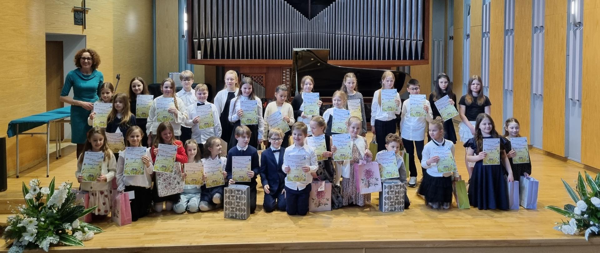 Zdjęcie przedstawia grupę dzieci stojących na scenie w sali koncertowej, najprawdopodobniej po zakończeniu występu lub konkursu. Uczestnicy: Na scenie znajduje się trzynaścioro dzieci w strojach galowych, które trzymają w rękach pamiątkowe dyplomy oraz kolorowe torebki z upominkami. Po lewej stronie widoczna jest również osoba dorosła, prawdopodobnie nauczycielka lub organizatorka, trzymająca mikrofon. Tło: Centralnym punktem tła są imponujące, nowoczesne organy o geometrycznym kształcie litery „V”, przed którymi stoi czarny fortepian skrzydłowy. Sceneria: Scena jest oświetlona jasnym światłem punktowym, a po jej obu stronach znajdują się dekoracje z białych kwiatów. W dolnej części zdjęcia widoczne są tyły głów publiczności siedzącej w niebieskich fotelach, w tym jedna osoba fotografująca wydarzenie telefonem. Całość ma uroczysty i radosny charakter, dokumentujący sukces młodych artystów.