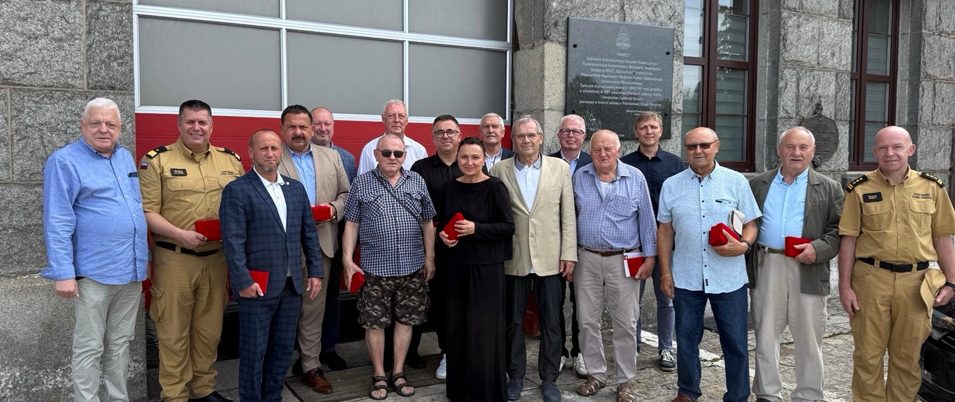 Zdjęcie przedstawia grupę 17 osób pozujących do wspólnej fotografii na zewnątrz budynku o szarej, kamiennej elewacji.
Otoczenie: Po lewej stronie widoczna jest duża, czerwona brama wjazdowa (garażowa) z oszklonymi panelami. Po prawej stronie na ścianie wisi ciemna pamiątkowa tablica z napisami oraz okna z kratami. Uczestnicy stoją na kostce brukowej.
Osoby na zdjęciu: W grupie przeważają mężczyźni w średnim i starszym wieku w strojach wizytowych oraz codziennych (garnitury, koszule, polówki). Jedyna kobieta stoi w środku, ubrana na czarno.
Strażacy: Na obu skrajach grupy stoją mężczyźni w piaskowych mundurach służbowych Państwowej Straży Pożarnej (po lewej jeden, po prawej drugi).
Detale: Większość osób w przednim rzędzie trzyma w rękach małe, czerwone, prostokątne pudełka na odznaczenia lub medale.