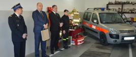 Przekazanie samochodu w OSP Jutrosin. Garaż w remizie OSP Jutrosin. Na zdjęciu widoczni pozostali goście: komendant PSP, minister Dziedziczak i burmistrz oraz członek MDP. Obok nich stolik ze sprzętem oraz przekazywany samochód.