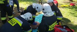 Manewry ratownicze. Teren byłego poligonu wojskowego w Sierakowie. Drużyna z Ochotniczej Straży Pożarnej w Roszkowie realizuje zadania na pierwszym stanowisku, gdzie doszło do targnięcia się młodego mężczyzny na swoje życie. Czterech druhów, klęcząc przy manekinie, prowadzi resuscytację krążeniowo-oddechową. Na tle drzew i zieleni pozoranci. Obok ratowników znajduje się torba ze sprzętem medycznym oraz nosze typu deska.