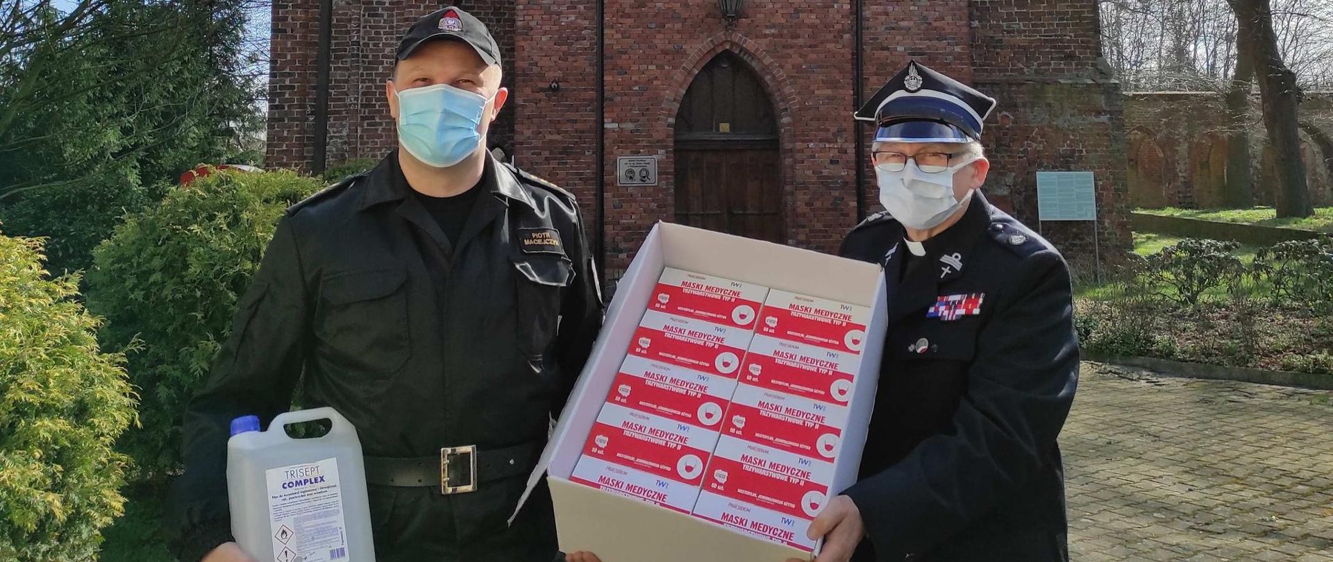 Na zdjęciu patrzący na wprost Komendant Powiatowy Państwowej Straży Pożarnej st. kpt Piotr Maciejczyk w czarnym mundurze trzymający w prawej ręce przeźroczysty pojemnik z płynem. Po jego lewej stronie w mundurze strażackim stoi proboszcz Parafii Rzymskokatolickiej pod wezwaniem Świętych Apostołów Piotra i Pawła ksiądz Waldemar Szczurowski. Obie osoby z twarzami w maseczkach trzymają pudło, w którego wnętrzu widoczne są czerwone etykiety opakowań. W tle zdjęcia ceglane mury budynku kościoła i tereny zielone.