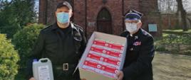 Na zdjęciu patrzący na wprost Komendant Powiatowy Państwowej Straży Pożarnej st. kpt Piotr Maciejczyk w czarnym mundurze trzymający w prawej ręce przeźroczysty pojemnik z płynem. Po jego lewej stronie w mundurze strażackim stoi proboszcz Parafii Rzymskokatolickiej pod wezwaniem Świętych Apostołów Piotra i Pawła ksiądz Waldemar Szczurowski. Obie osoby z twarzami w maseczkach trzymają pudło, w którego wnętrzu widoczne są czerwone etykiety opakowań. W tle zdjęcia ceglane mury budynku kościoła i tereny zielone.