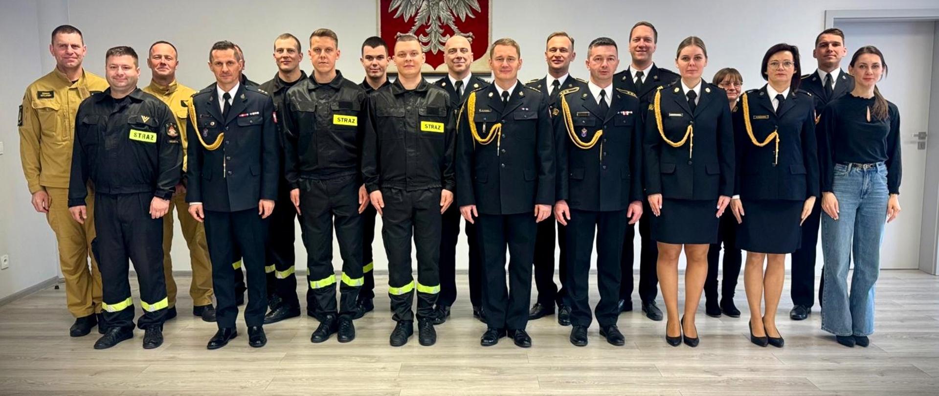 Na zdjęciu widzimy grupę strażaków zgromadzonych w sali. Wszyscy stoją wyprostowani.
W tle znajduje się godło Polski – biały orzeł na czerwonym tle, co sugeruje oficjalny charakter wydarzenia. Zdjęcie wykonane jest w jasnym pomieszczeniu z nowoczesną podłogą i białymi ścianami.