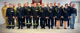 Na zdjęciu widzimy grupę strażaków zgromadzonych w sali. Wszyscy stoją wyprostowani.
W tle znajduje się godło Polski – biały orzeł na czerwonym tle, co sugeruje oficjalny charakter wydarzenia. Zdjęcie wykonane jest w jasnym pomieszczeniu z nowoczesną podłogą i białymi ścianami.