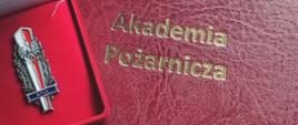 Odznaka Akademii Pożarniczej w czerwonym etui na tle teczki z logo i nazwą Akademii Pożarniczej.