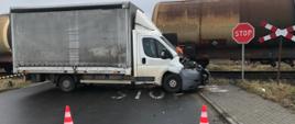 Na zdjęciu widać samochód dostawczy z uszkodzonym przodem. Pojazd stoi w poprzek jezdni natomiast za nim widać skład kolejowy z wagonami typu cysterna