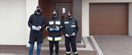 Zdjęcie przedstawia dwóch strażaków OSP, którzy wręczają mężczyźnie ulotkę dotyczącą szczepień przeciwko COVID-19. Zdjęcie wykonane jest na tle budynku mieszkalnego, widać drzwi wejściowe do budynku oraz drzwi garażowe.