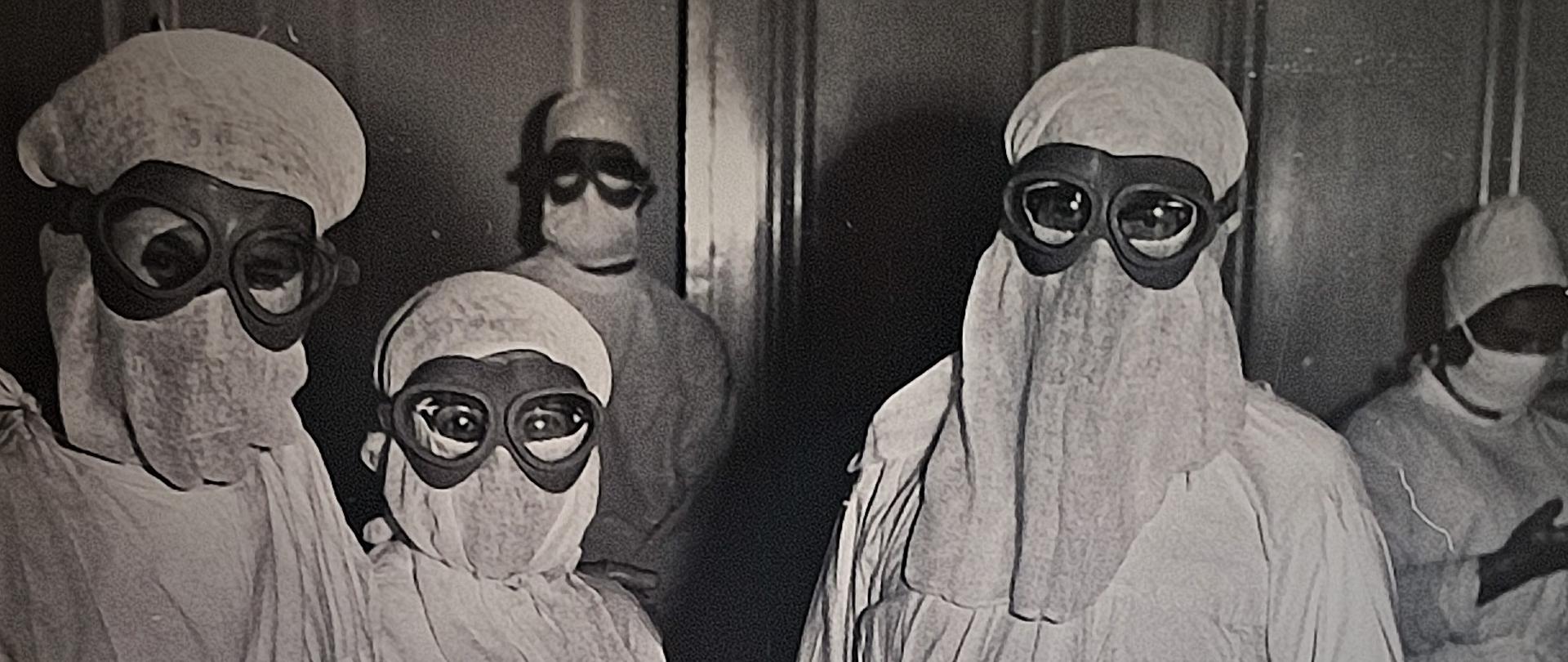 Troje lekarzy w kompletnych strojach ochronnych. Osoby ubrane w fartuchy, czepki, maseczki, rękawice gumowe oraz gogle ochronne na oczy.