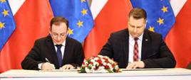 Przy białym stole siedzi minister Czarnek i mężczyzna w czarnym garniturze i okularach, obaj podpisują leżące na stole dokumenty, przed nimi mały bukiet biało-czerwonych kwiatów, za nimi rząd flag Polski i UE.