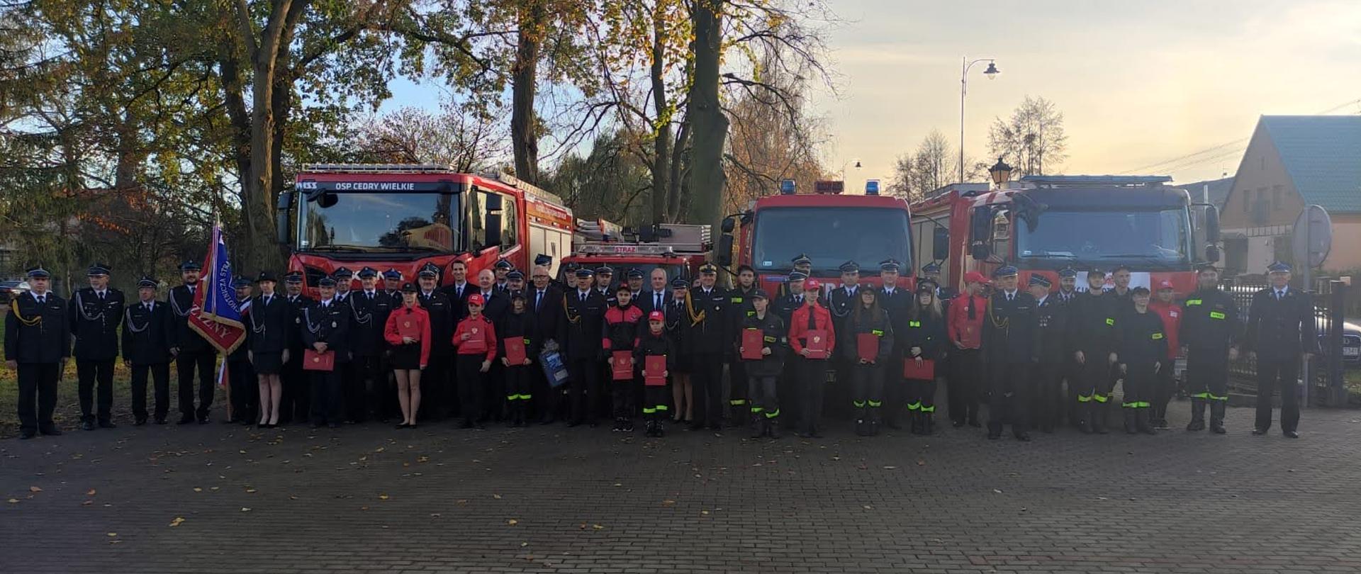 Zaproszeni goście, strażacy Państwowej Straży Pożarnej oraz ochotniczych straży pożarnych stoją obok siebie za nimi ustawione są samochody gaśnicze.