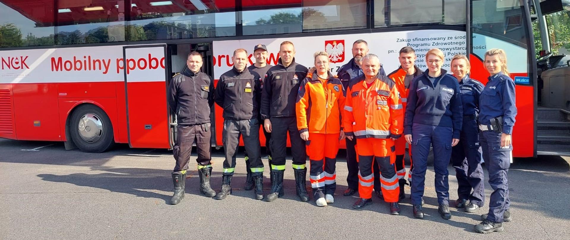 Obraz przedstawia strażaków, policjantów i pracowników pogotowia ratunkowego stojących na tle mobilnego punku poboru krwi.