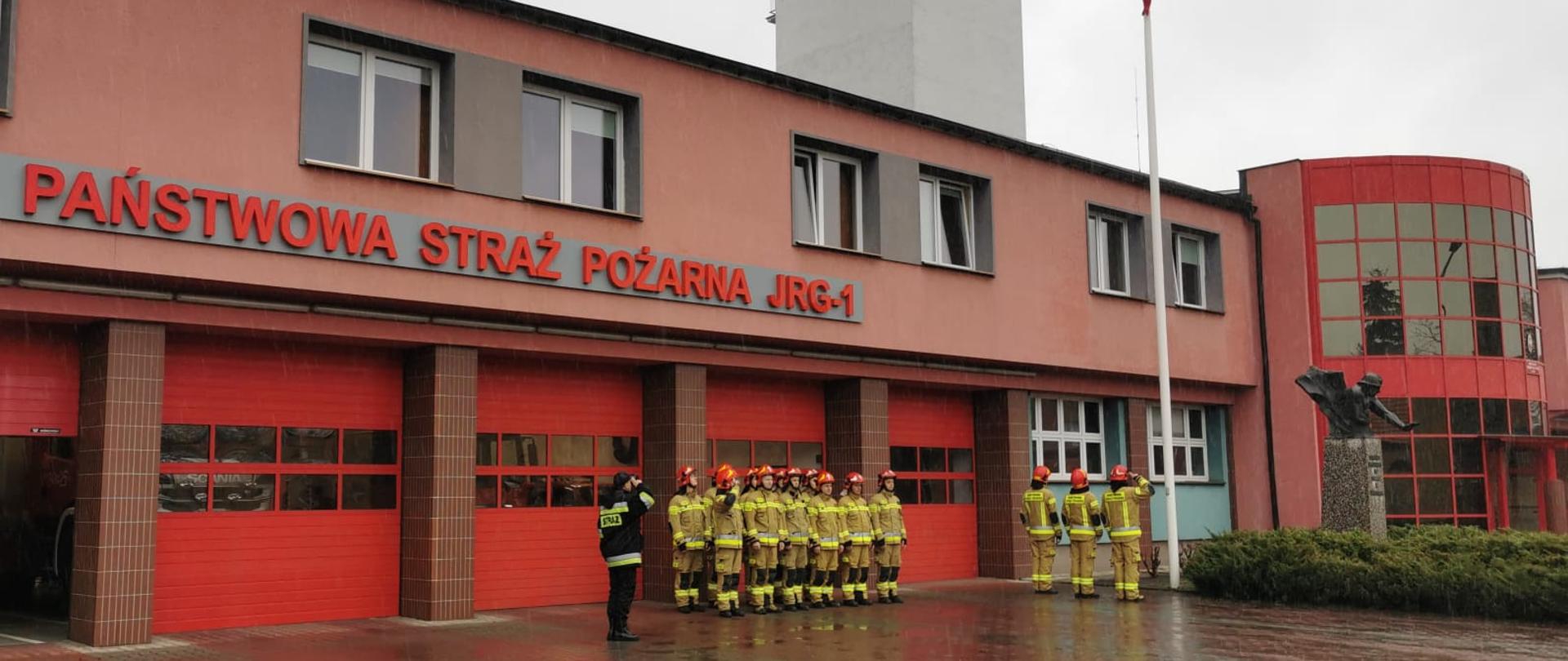 Na zdjęciu widać poczet flagowy. Ubrany jest w ubrania specjalne koloru piaskowego oraz czerwone hełmy. Jeden ze strażaków niesie na rękach flagę państwową w kolorze biało-czerwonym. W tle widać budynek Jednostki Ratowniczo-Gaśniczej nr 1. Strażacy idą po kostce brukowej.
