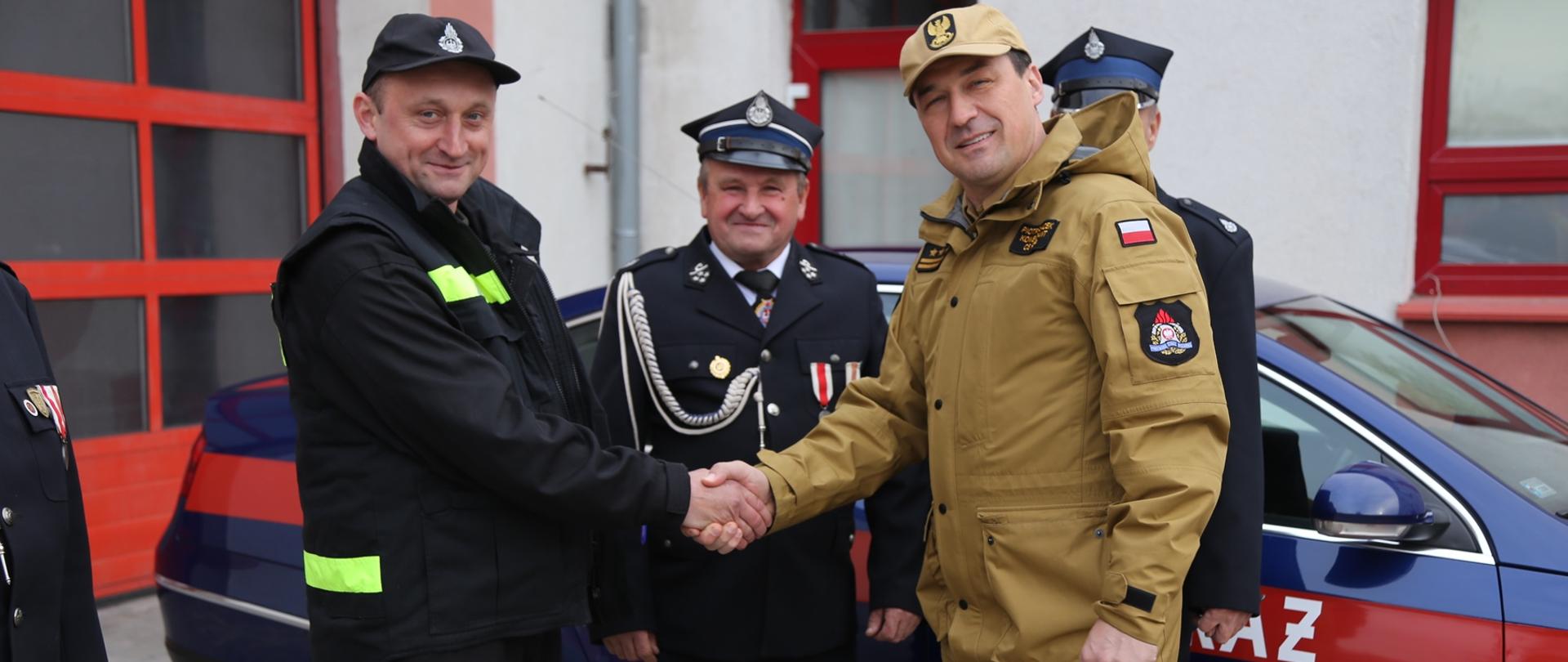 Na zdjęciu Komendant Centralnej Szkoły PSP w uścisku dłoni z druhem OSP, w głębi dwóch druhów OSP oraz przekazywany samochód VW Passat