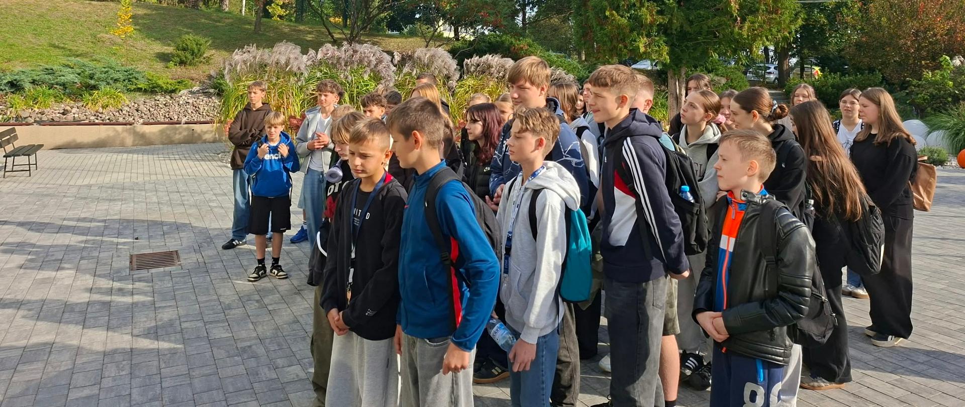 Grupa młodzieży stojąca na placu przed szkołą w otoczeniu zieleni i drzew w ZSCKR Samostrzel.
