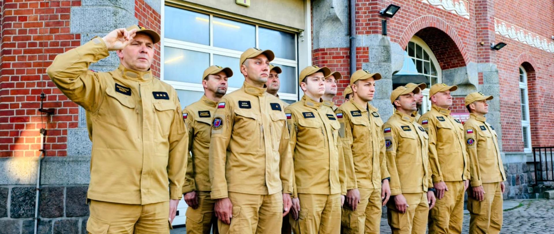 Na zdjęciu pododdział strażaków w czasie zbiórki, w tle widoczny ceglano kamienny straży pożarnej