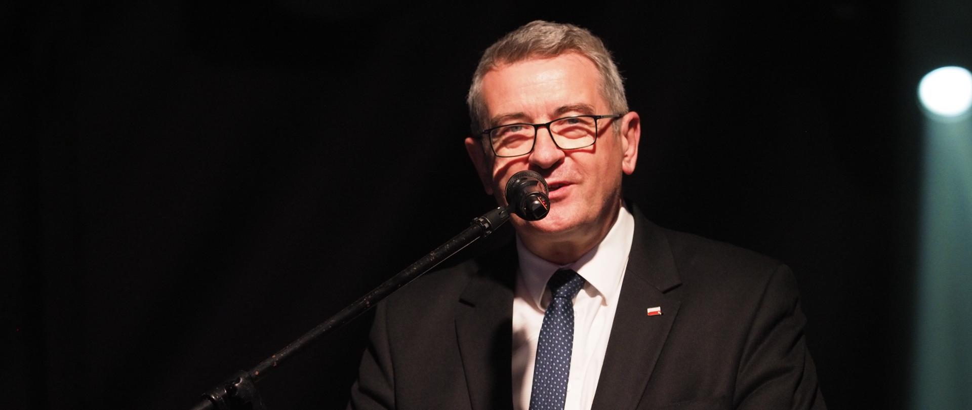 Wojciech Murdzek przemawia podczas gali Gwiazdy biznesu do mikrofonu. ubrany w czarny garnitur i białą koszulę. widoczny na czarnym tle.