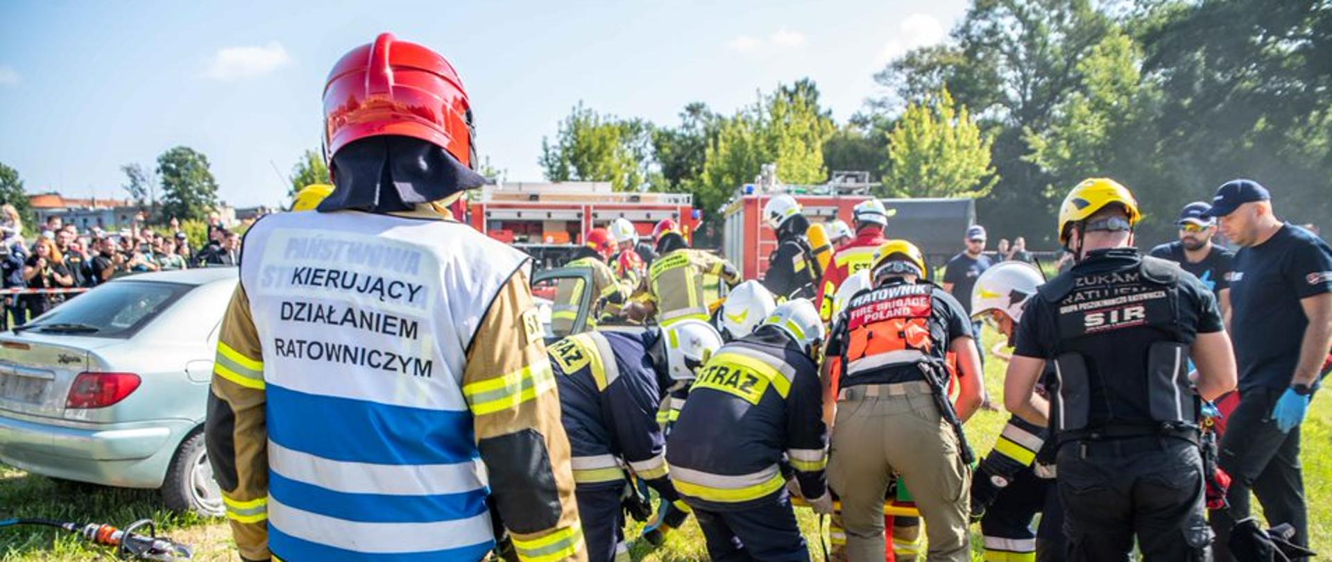strażacy i inne służby przenoszą na desce ortopedycznej osobę poszkodowaną, przd nimi stoi strażak w hełmie i kamizelce z napisem kierujący działaniem ratowniczym, w tle pojazdy pożarnicze
