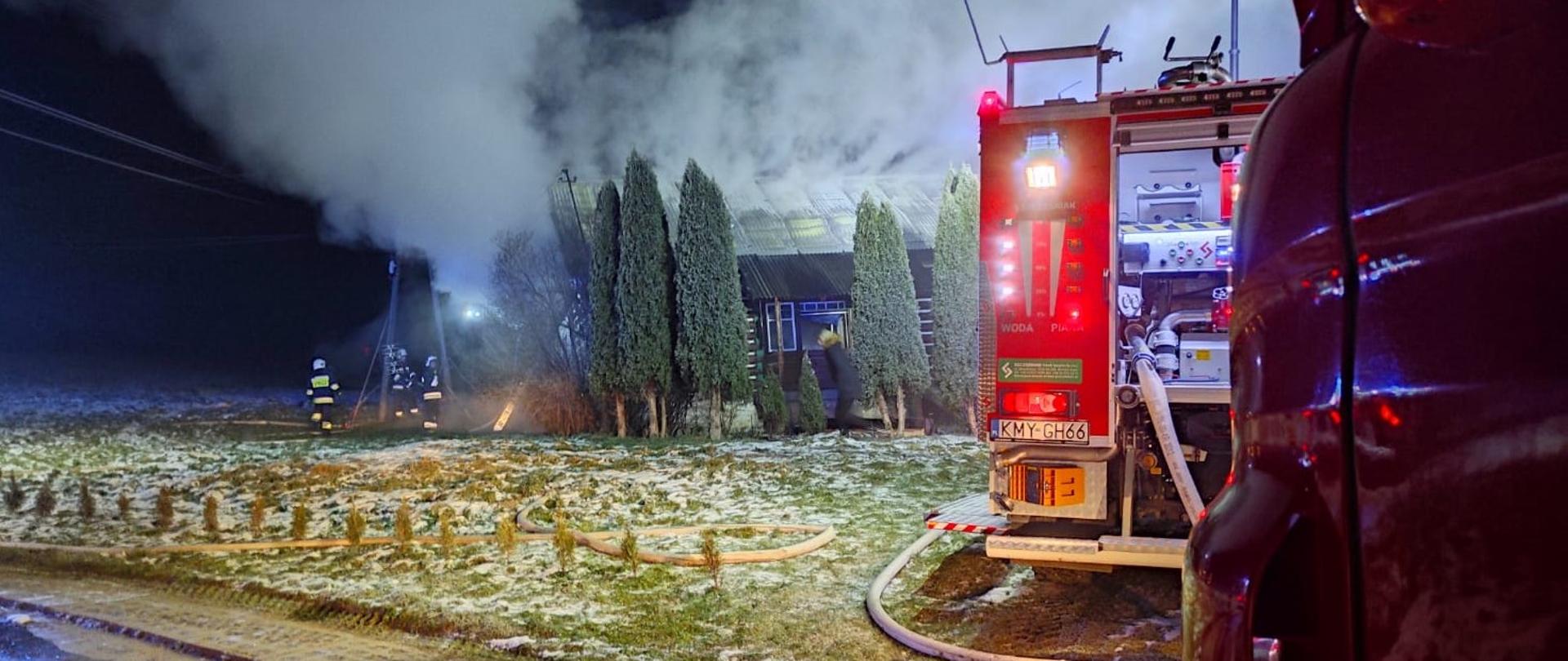 Zdjęcie przedstawia pożar budynku mieszkalnego mającego miejsce w porze nocnej. Z prawej strony zdjęcia widoczne są samochody strażackie. W oddali widoczna grupa strażaków prowadząca działania gaśnicze. Z obiektu wydobywają się duże kłęby dymu.