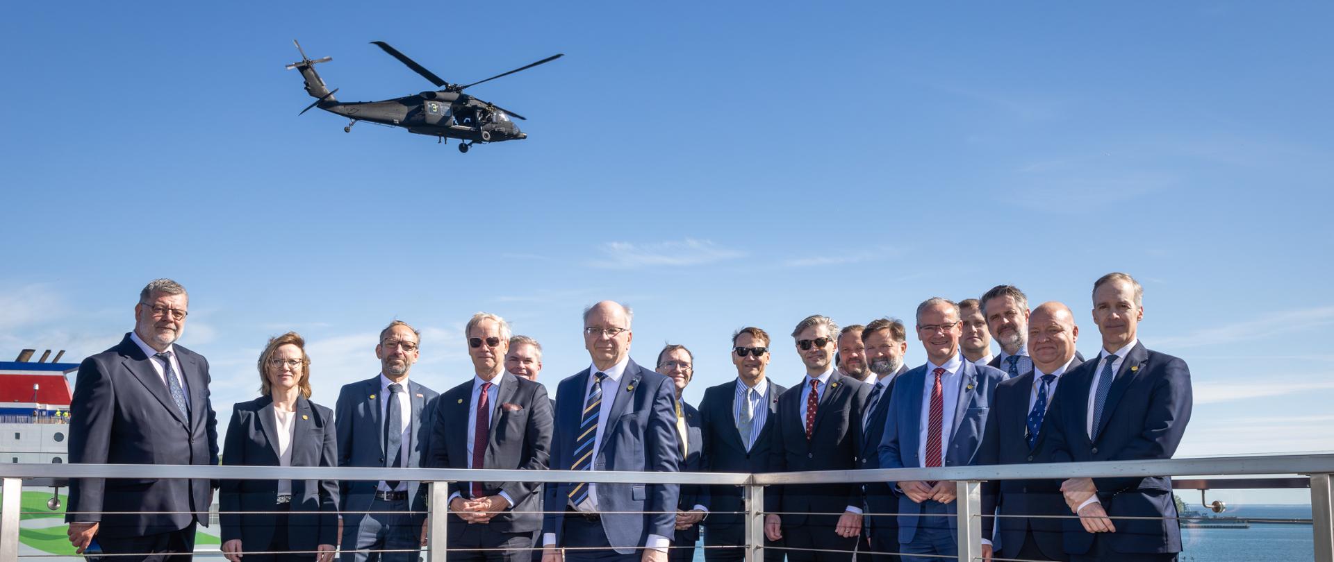 Na zdjęciu jest grupa osób w garniturach. Za nimi jest akwen wodny. Nad nimi jest śmigłowiec. 