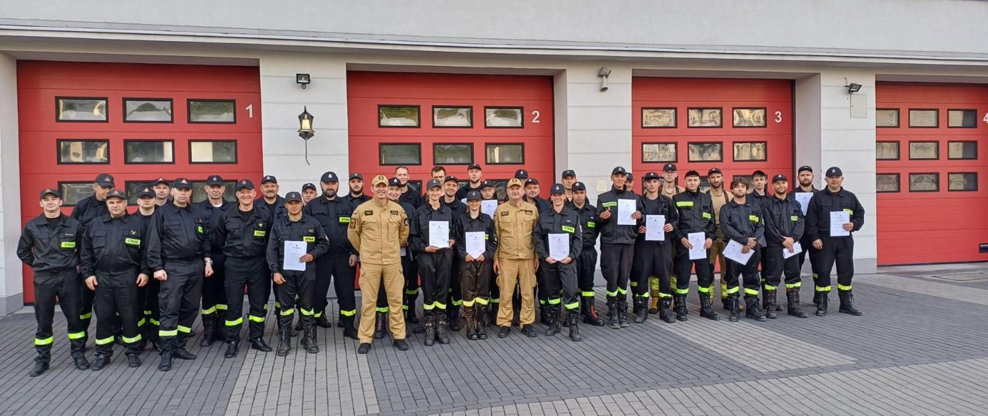 Na zdjęciu widać grupę strażaków. Strażacy są ubrani w czarne mundury. Dwóch strażaków stojących po środku ma żółte mundury. Strażacy stoją na tle budynku komendy. W tle widać trzy bramy garażowe w kolorze czerwonym. Nad bramami widać okna. Budynek jest szarego koloru. Strażacy stoją na placu komendy. Na ziemi widać kostkę brukową koloru szarego.