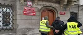 Zatrzymany prowadzony przez funkcjonariuszy w kamizelkach z napisem KAS przed budynkiem Prokuratury Regionalnej w Warszawie.
