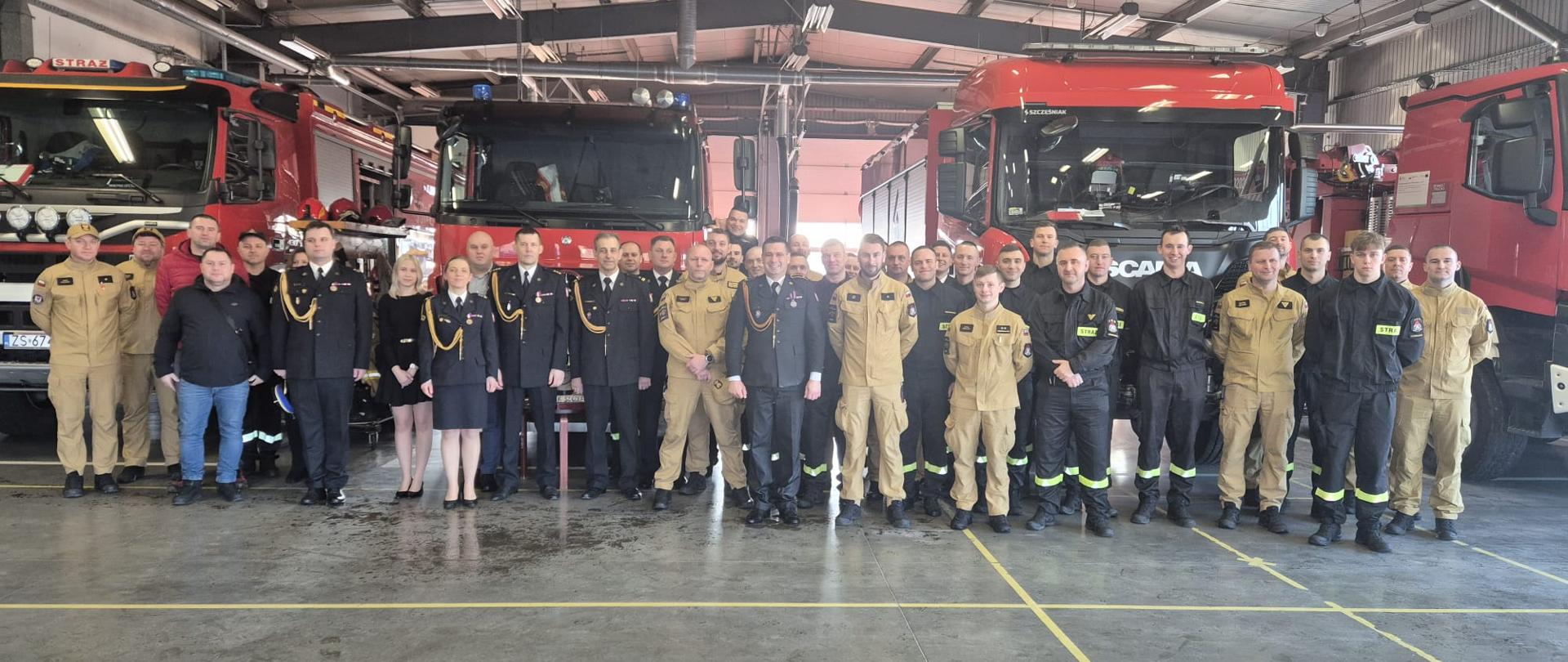 Zdjęcie grupowe kilkudziesięciu strażaków i strażaczek pozujących wewnątrz garażu remizy. W tle widoczne są trzy duże, czerwone wozy strażackie. Zgromadzeni noszą różne rodzaje umundurowania: od ciemnych mundurów galowych ze sznurami funkcyjnymi i medalami, przez piaskowe mundury służbowe, aż po czarne mundury koszorowe z żółtymi elementami odblaskowymi i napisem „STRAŻ”.