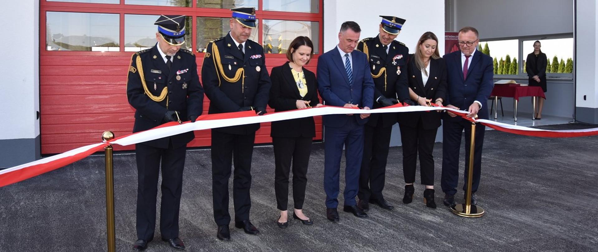Na zdjęciu widzimy moment przecięcia wstęgi przez zaproszonych gości, a także kierownictwo komendy powiatowej i wojewódzkiej. Za przecinającymi wstęgę osobami znajduje się rozbudowana część obiektu strażnicy.