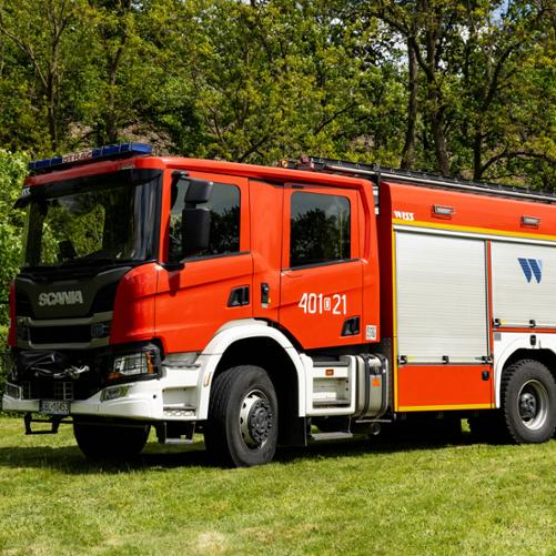 Średni samochód ratowniczo-gaśniczy marki Scania P360 w kolorze czerwonym z białymi nadkolami i zderzakiem. Na drzwiach kabiny znajduje się numer operacyjny „401[D]21”, a na bocznych, srebrnych żaluzjach widoczne jest niebieskie logo „WISS”. Wóz zaparkowany jest na trawie przed ścianą lasu.