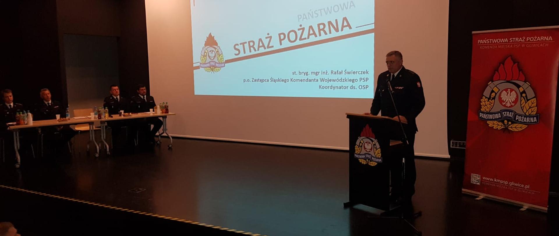 Na scenie za mównicą przemawia p.o. zastępcy Śląskiego Komendanta Wojewódzkiego PSP st. bryg. Rafał Świerczek , z boku sceny przy stolikach oraz pod sceną widać siedzące audytorium 