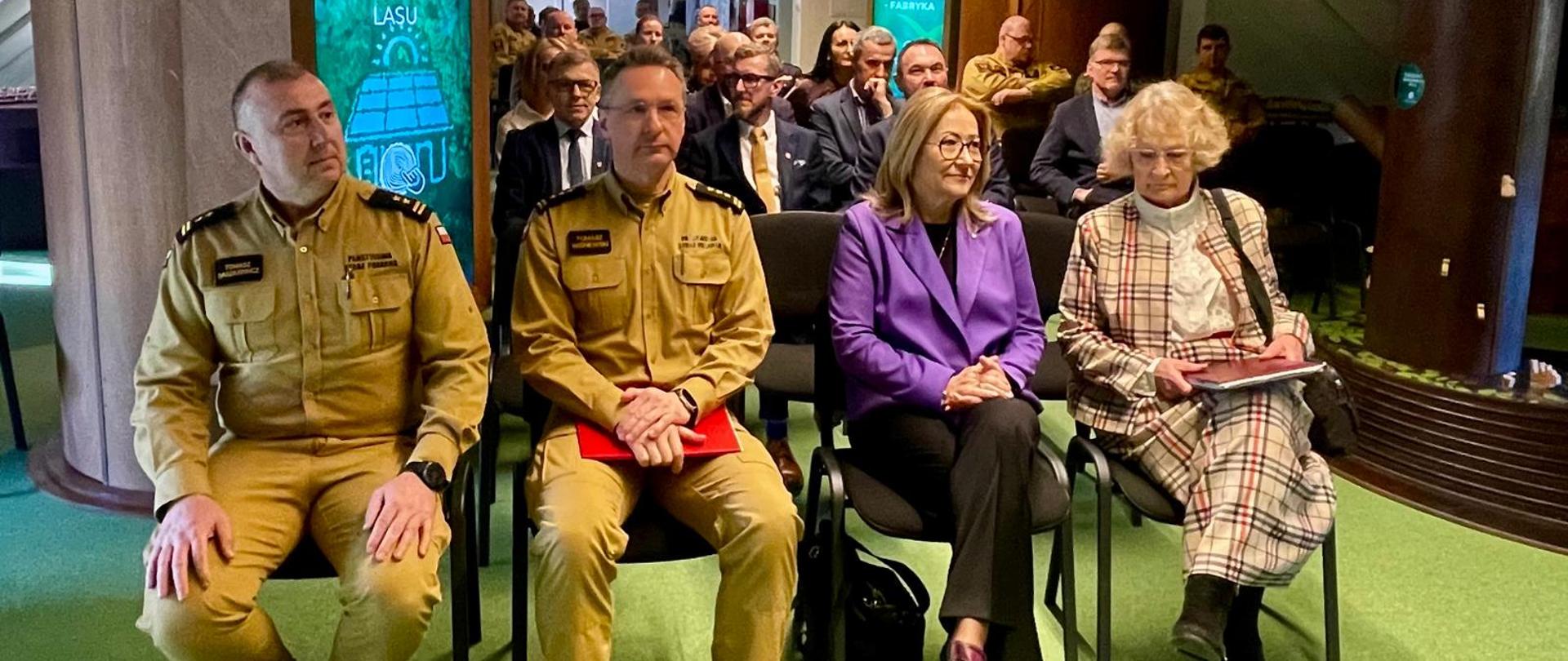 Na pierwszym planie siedzą cztery osoby podczas spotkania – dwóch mężczyzn w mundurach oraz dwie kobiety w cywilnych strojach. W tle widać uczestników wydarzenia siedzących w rzędach krzeseł w sali konferencyjnej.