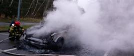 Zdjęcie przedstawia strażaka dogaszającego samochód osobowy. Strażak pochyla się przy nadkole pojazdu i podaje środek gaśniczy za pomocą linii wężowej. 