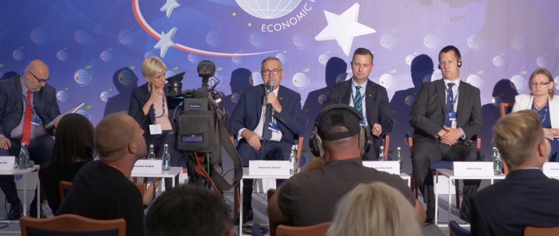 Zdjęcie debatujących. Wszyscy siedzą. W środku wiceminister Szwed. W ręku trzyma mikrofon. Przemawia do zgromadzonych.