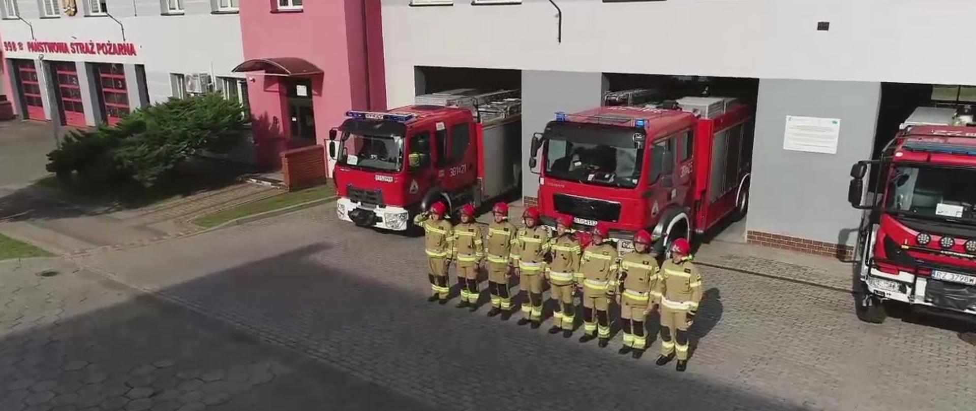 Przed budynkiem strażnicy w szeregu stoją strażacy umundurowani w ubrania bojowe oraz hełmy. Za nimi stoją samochody pożarnicze. 