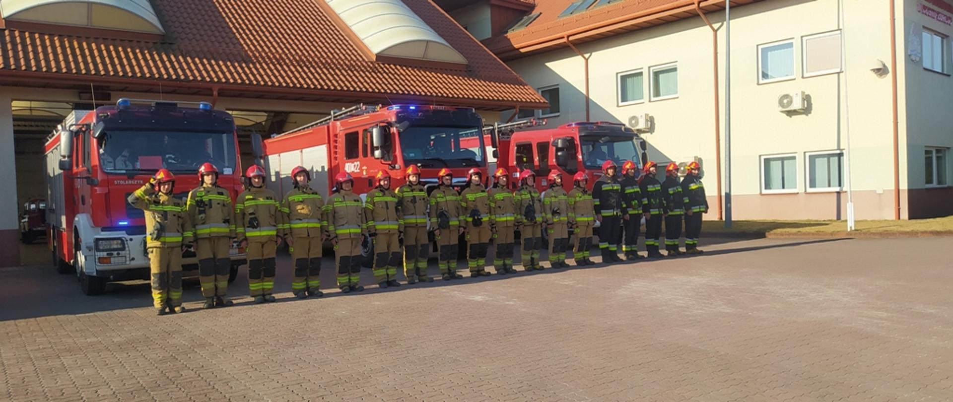 Zdjęcie przedstawia pododdział strażaków w umundurowaniu specjalnym, stojący w rzędzie przed pojazdami pożarniczymi. Strażak stojący na czele pododdziału salutuje. Pojazdy pożarnicze mają włączone światła blyskowe