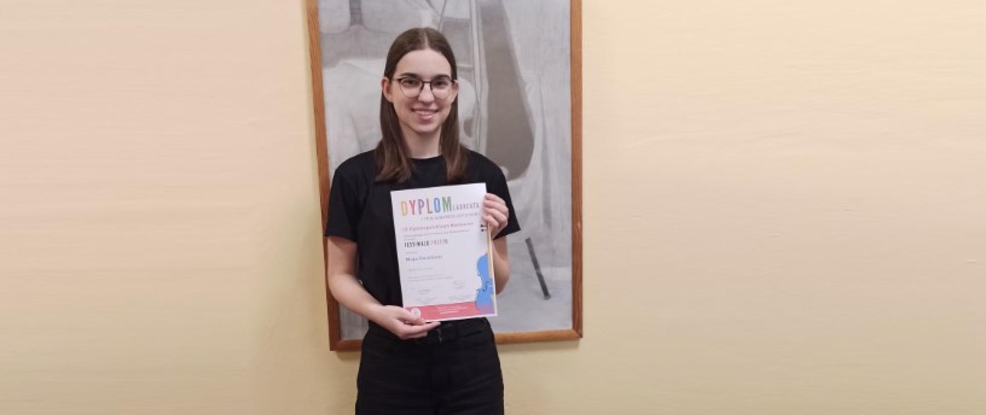 Ciemnowłosa dziewczyna w okularach trzyma w rękach dyplom. Za nią czarno - biały obraz - martwa natura ze skrzypcami.