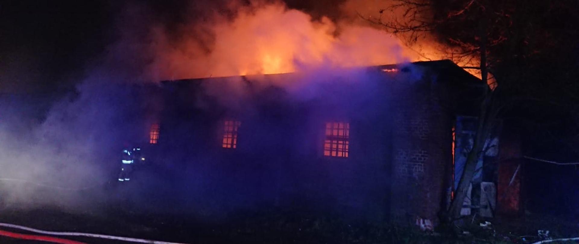 Jest ciemno - noc. Widać stary budynek z cegły, magazynowy, objęty pożarem. Rozwinięte linie gaśnicze i strażacy przy oknie.