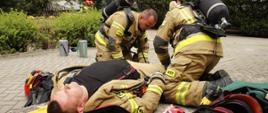 Na pierwszym planie znajduje się leżący na desce ratowniczej strażak pozorujący poszkodowanie. W dalszym planie widocznych jest dwóch strażaków z aparatami powietrznymi na plecach udzielający pomocy poszkodowanemu.