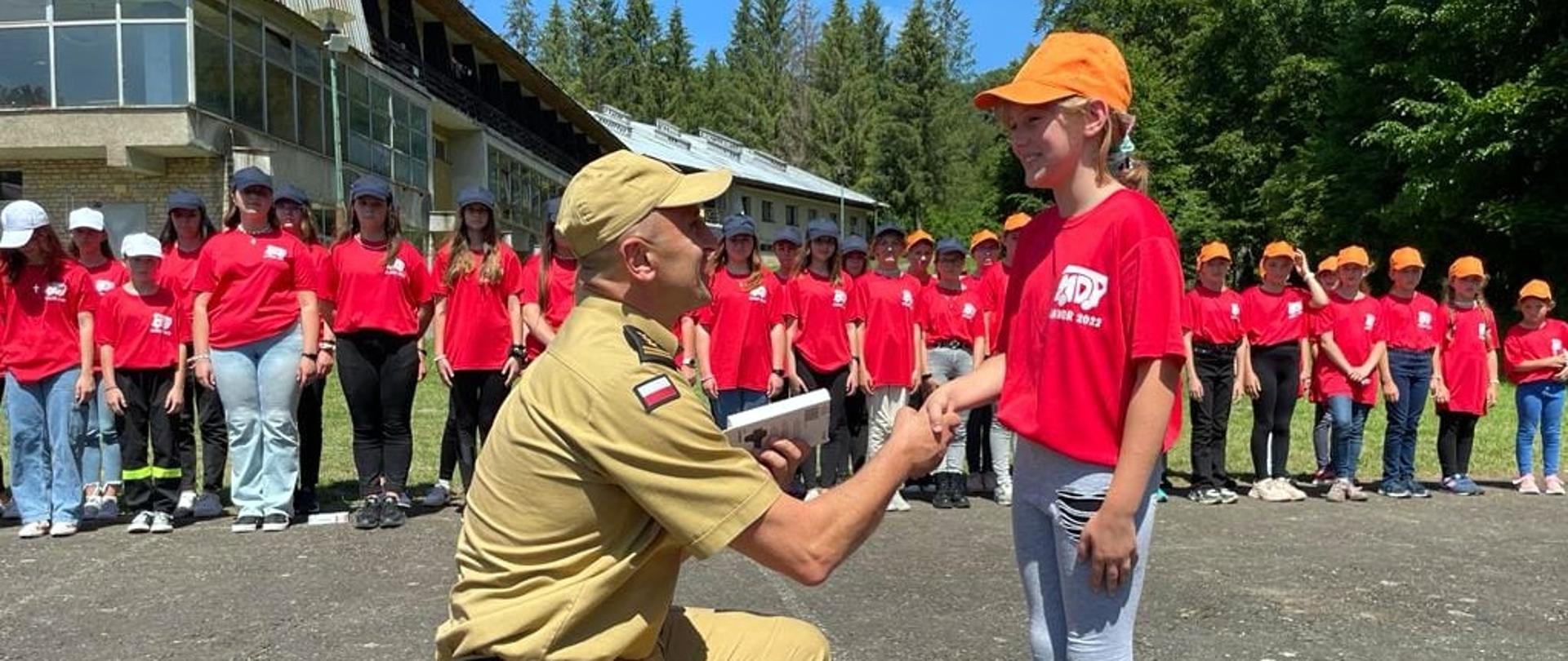 Zdjęcie zrobione na zewnątrz podczas uroczystej zbiórki Młodzieżowych Drużyn Pożarniczych. Oficer Państwowej Straży Pożarnej podaje dłoń dziewczęce z Młodzieżowej Drużyny Pożarniczej. W tle widać uczestników obozu zebranych na uroczystej zbiórce.
