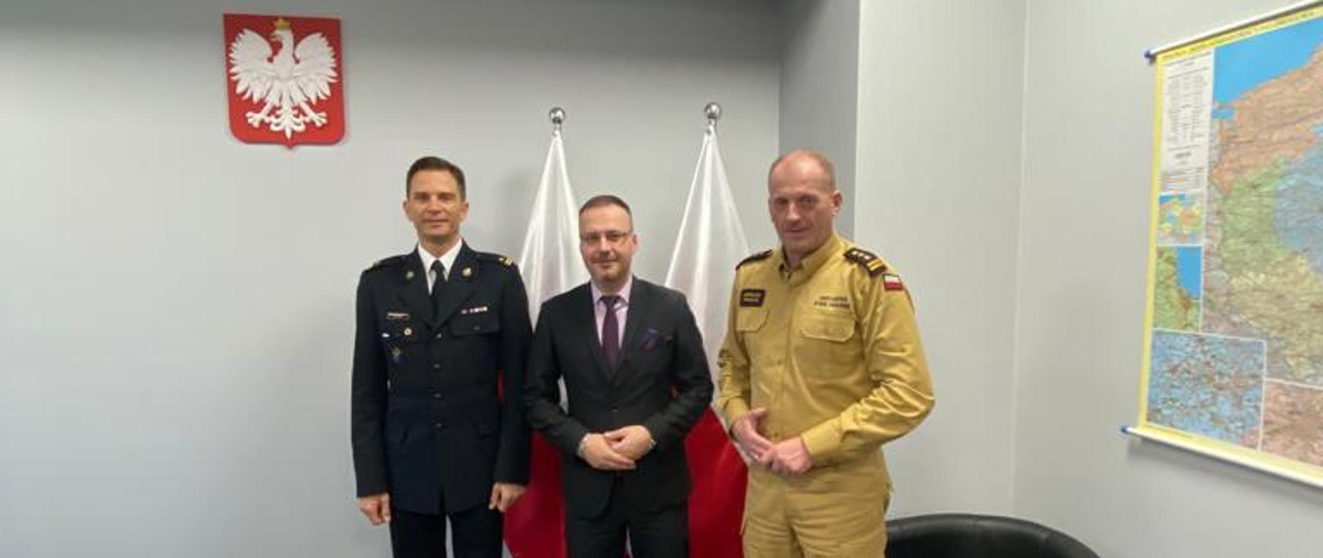 Na zdjęciu 3 mężczyzn stojacych w gabinecie. Na ścianie mapa Polski, godło. Wewnątrz biurko.