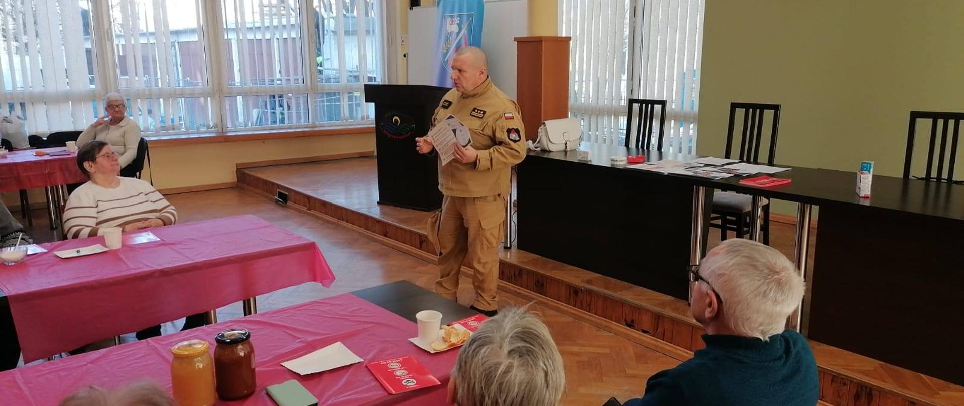 Duża sala wykładowa. Strażak prowadzi szkolenie i pokazuje czujkę czadu i dymu. Przy stołach siedzą słuchacze.