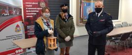 Dwie harcerki w mundurkach przekazują Komendantowi PSP w Brzezinach Światło Pokoju. Wszystkie osoby w maseczkach w pomieszczeniu KP PSP w Brzezinach. 